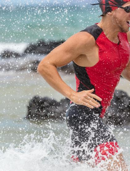 Atleta corre fuori dall'acqua in spiaggia, indossa costume rosso e occhiali.