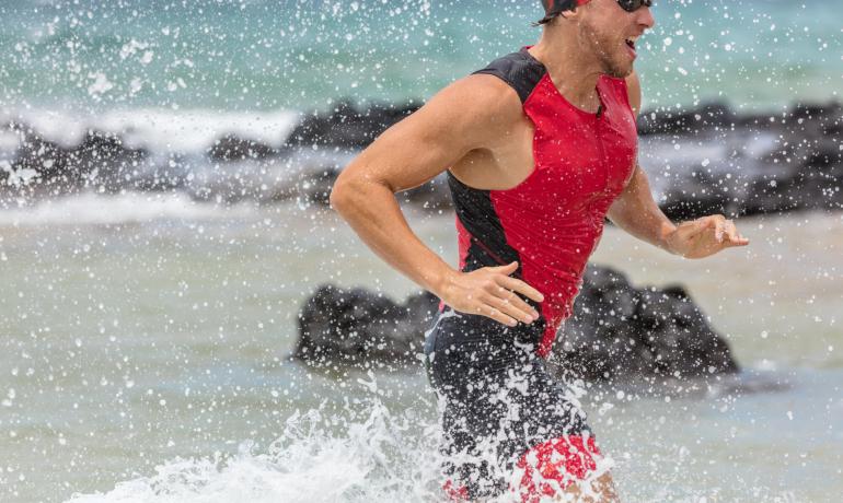 Atleta corre fuori dall'acqua in spiaggia, indossa costume rosso e occhiali.