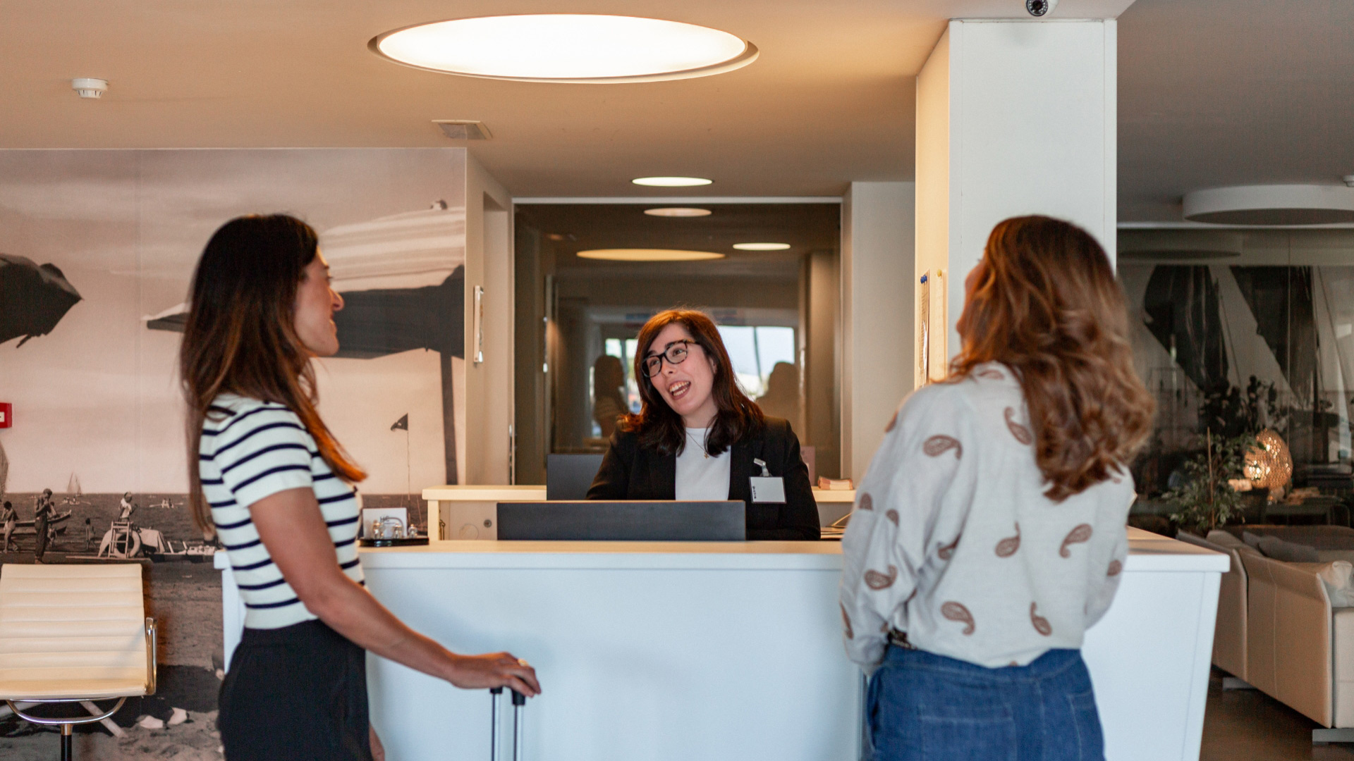 Reception di un hotel con due ospiti e una receptionist sorridente.