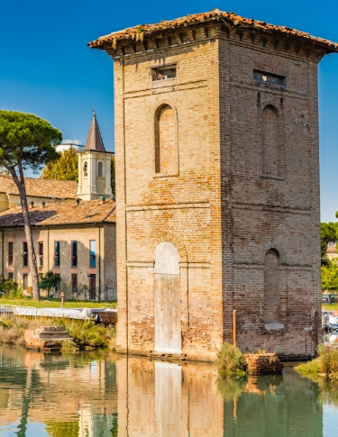 Torre in mattoni riflessa nell'acqua, con edifici storici e alberi sullo sfondo.