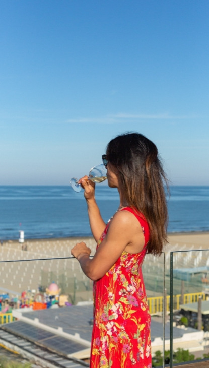 Donna in abito floreale beve vino su una terrazza con vista mare.