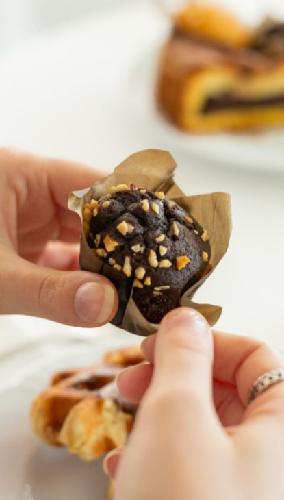 Mani che tengono un muffin al cioccolato con granella di nocciole su un tavolo.