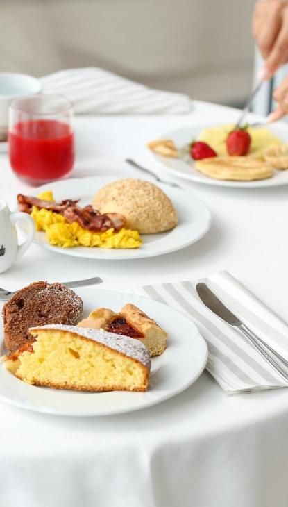 Colazione con dolci, uova, pancetta, bevande e frutta su tavola apparecchiata.