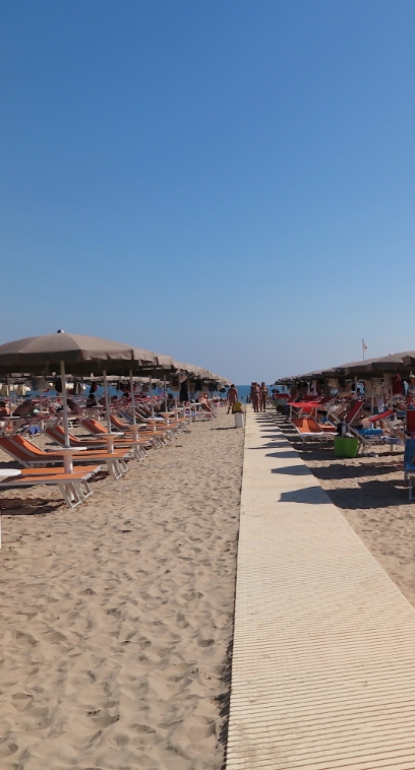 Spiaggia con lettini e ombrelloni, cielo sereno e passerella in legno.