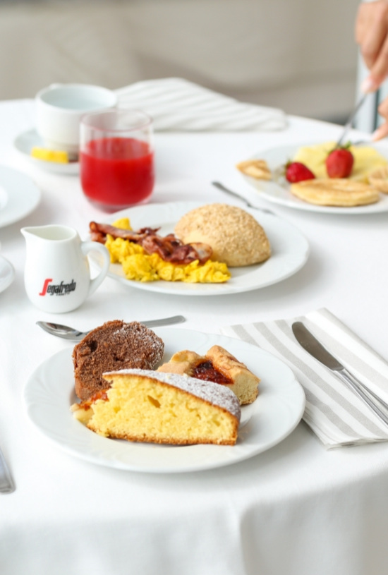 Colazione con dolci, uova, bacon, frutta, caffè e succo su tavola apparecchiata.